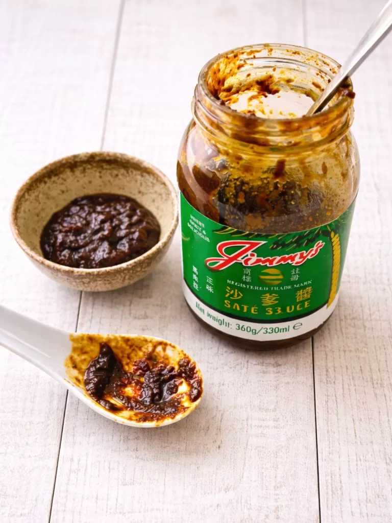 Open jar of Jimmy’s satay sauce on a white timber surface with a small bowl and spoonful of thick, dark satay sauce in natural light.