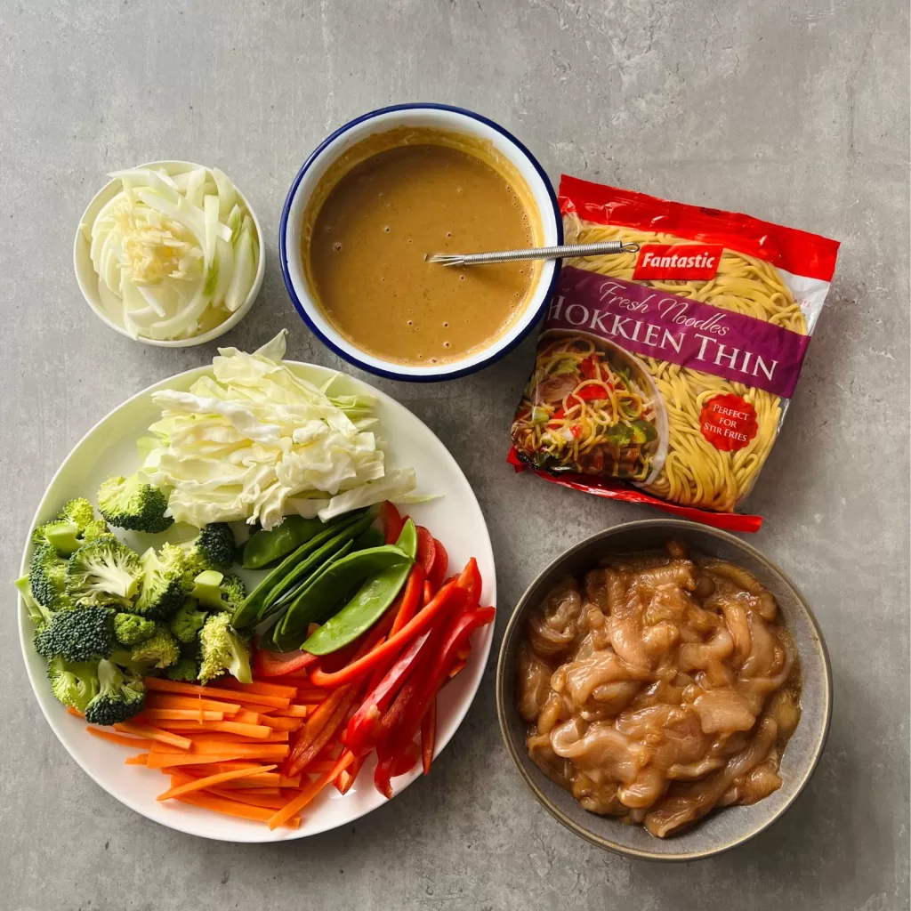 Overhead view of the prep ingredients for satay chicken noodles, including vegetables, marinated chicken, satay sauce and hokkien noodles.