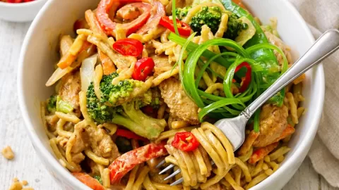 Creamy satay chicken noodles in a white bowl, tossed with tender chicken, broccoli, capsicum and carrots, topped with sliced red chilli and curled spring onion, with a fork twirling noodles, served on a white timber surface with lime wedges and satay sauce on the side.