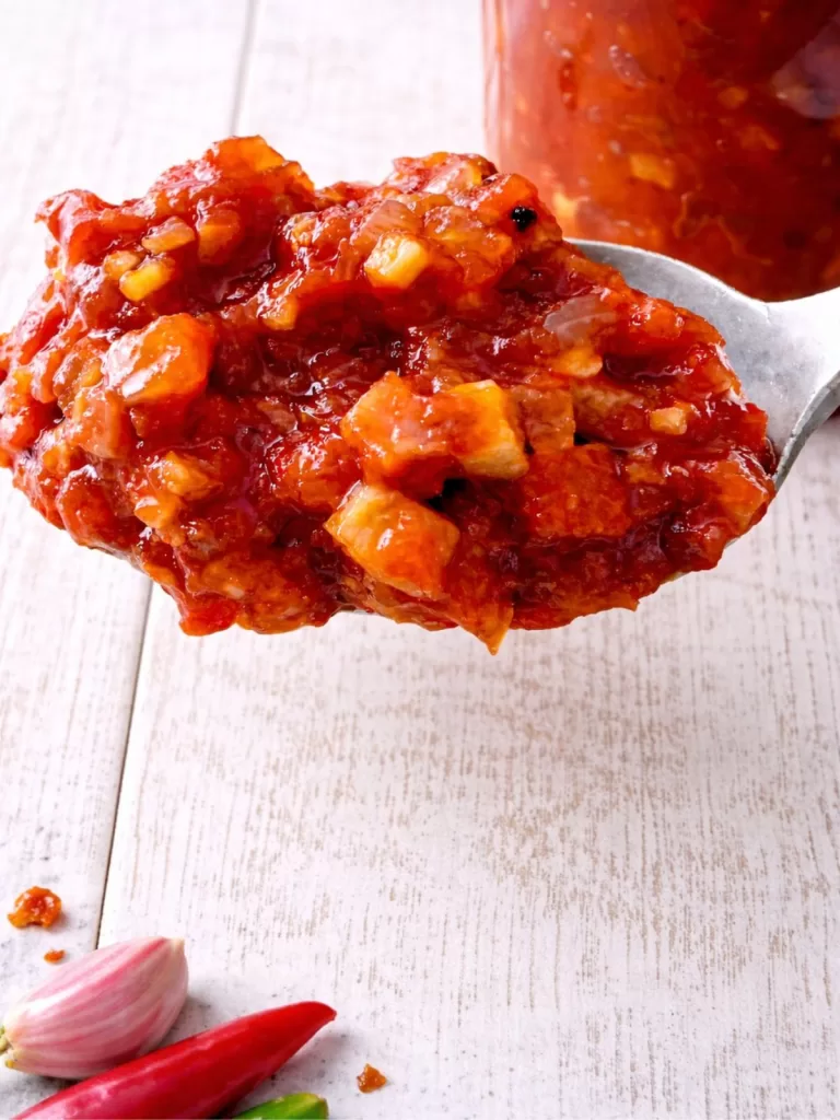 Heaped spoonful of tomato bacon chilli jam with visible bacon and tomato pieces, held above a jar on a white timber background in soft natural light.