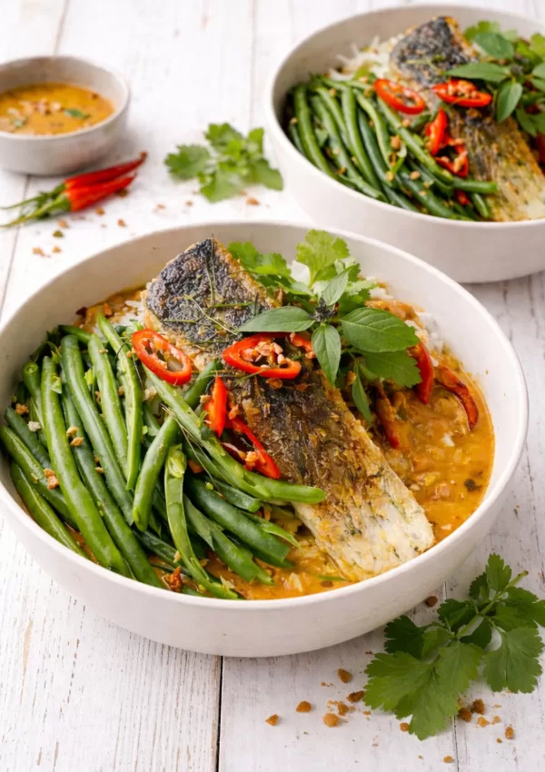 Crispy skin barramundi served over jasmine rice with Thai panang curry sauce, topped with green beans, red chilli, fried shallots and Thai basil and coriander in a white bowl.