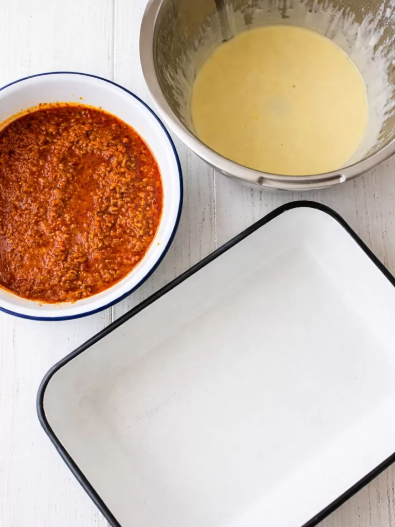 Top-down view of lasagna prep on a white timber surface, showing a bowl of meat sauce, a bowl of creamy béchamel, and an empty white baking dish in bright natural light.