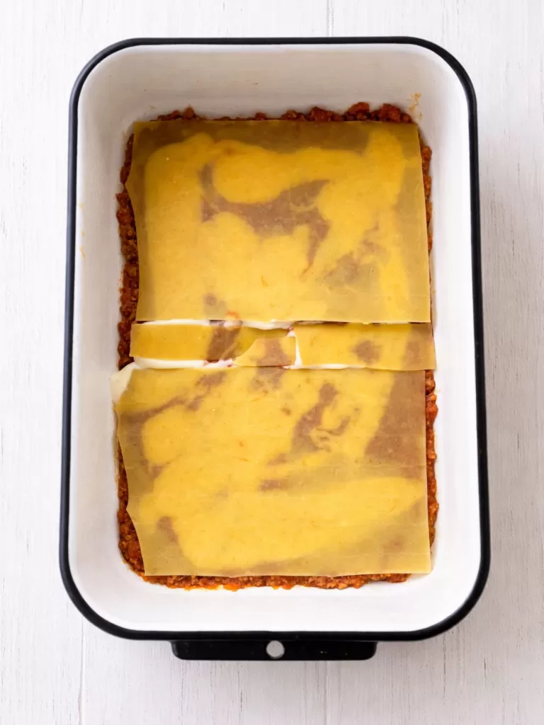Overhead view of a lasagna being assembled in a white baking dish, with a layer of meat sauce topped by pasta sheets and béchamel on a white timber surface in bright light.