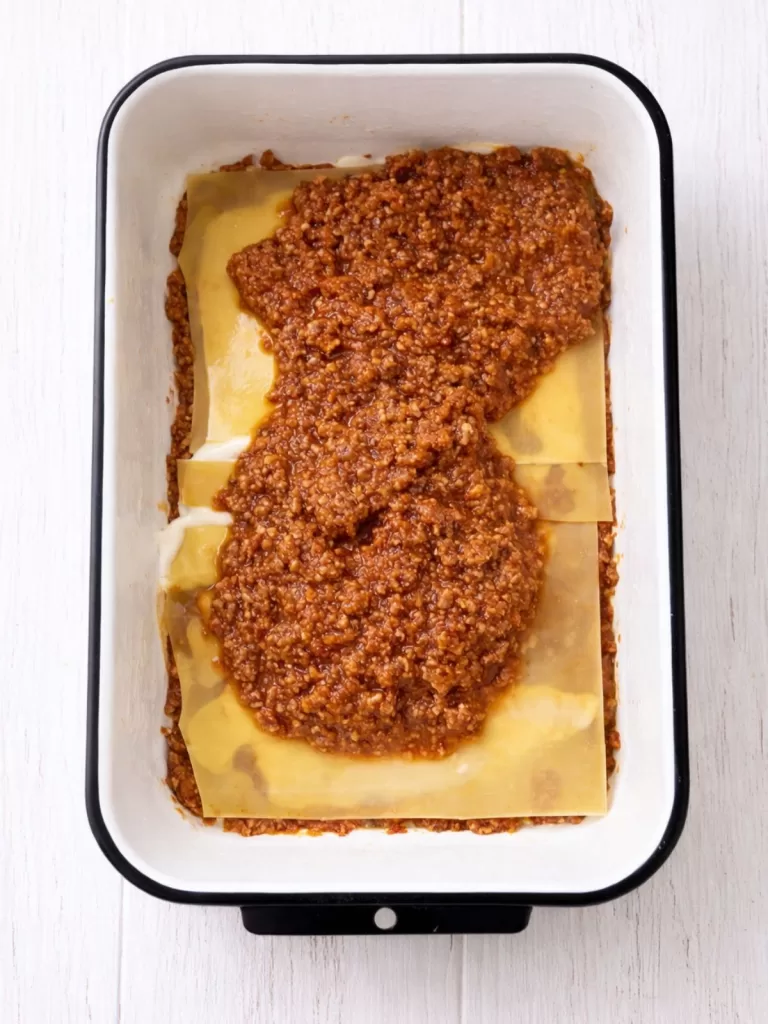 Overhead view of a lasagna being layered in a white baking dish, showing pasta sheets topped with béchamel and a thick layer of meat ragu on a white timber surface in bright light.