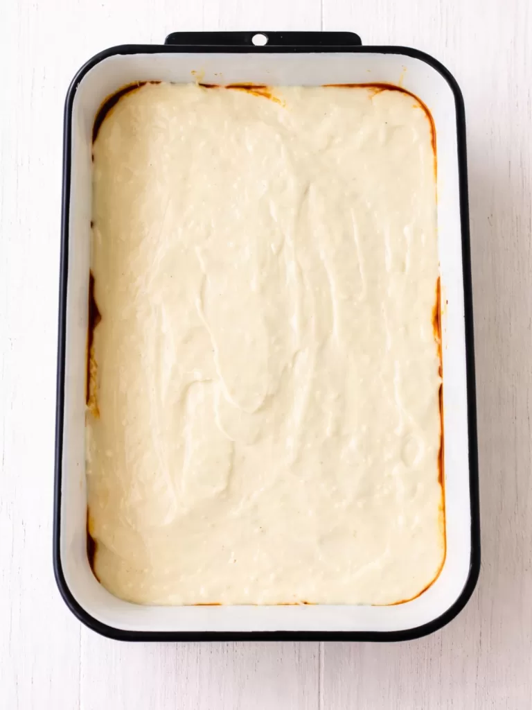 Overhead view of a white baking dish filled with lasagna topped with a smooth layer of béchamel sauce, set on a white timber surface in bright light.