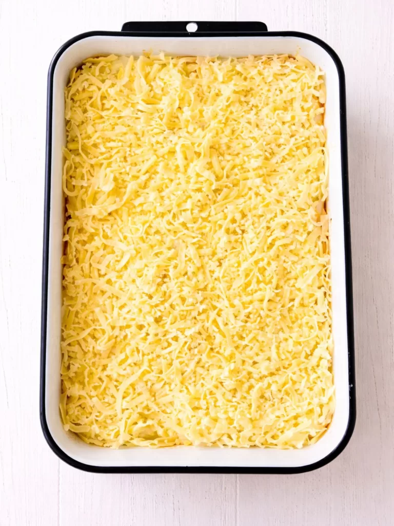 Overhead view of a white baking dish filled with uncooked lasagna topped with a thick layer of grated cheese, on a white timber surface in bright light.