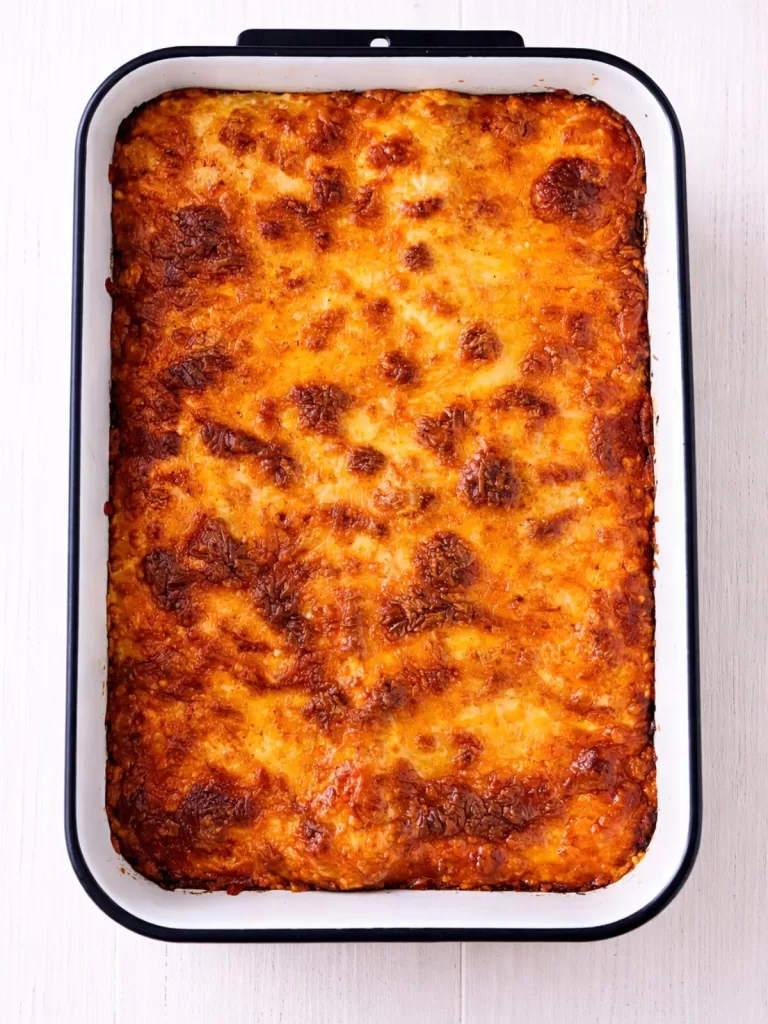 Overhead view of a baked lasagna in a white baking dish with a golden, bubbly cheese topping, set on a white timber surface in bright light.