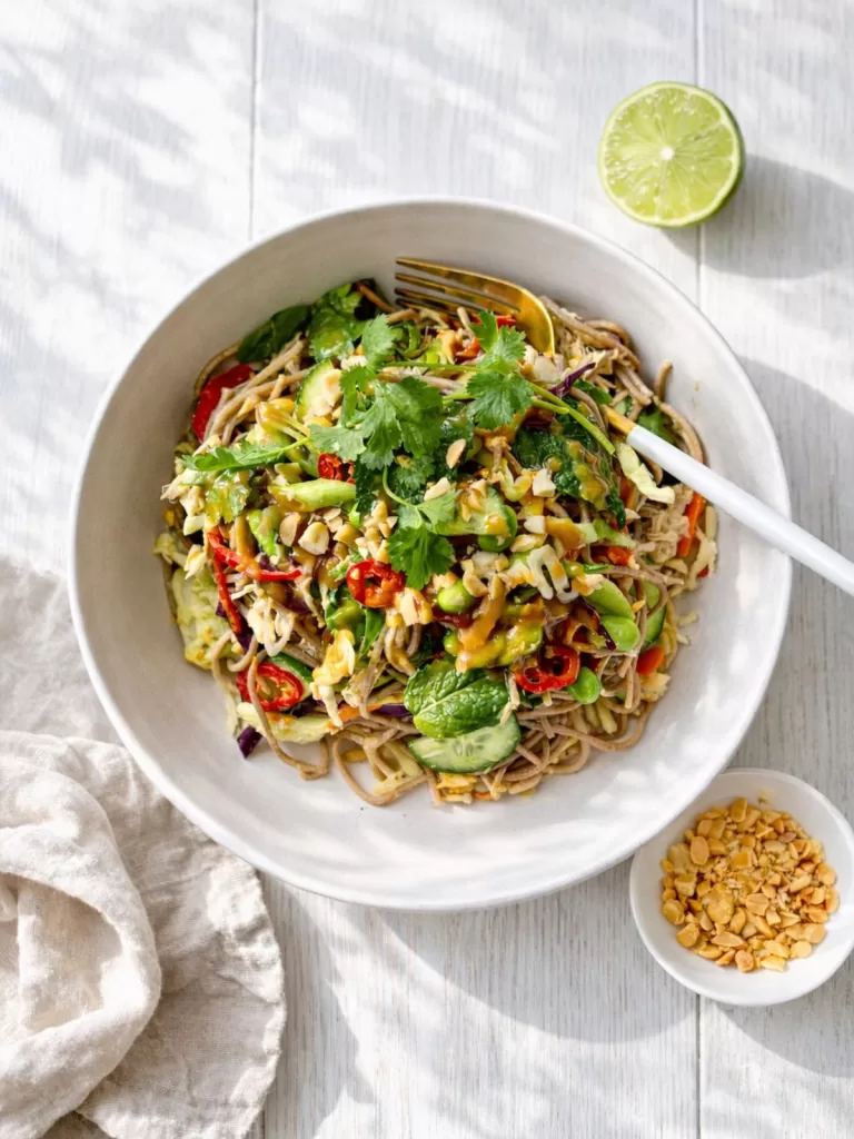 Poached chicken soba noodle salad with crunchy vegetables, fresh herbs and creamy peanut dressing in a white bowl on a light timber background.