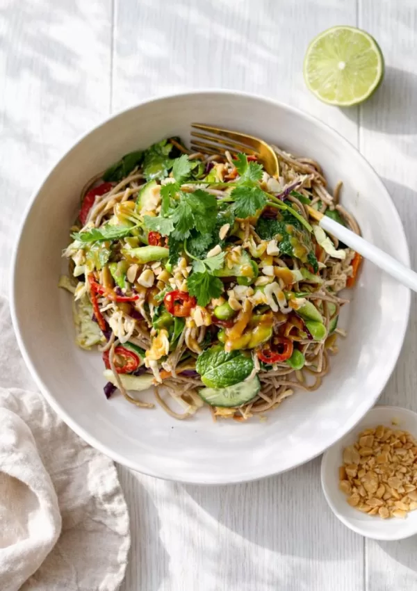 Poached chicken soba noodle salad with crunchy vegetables, fresh herbs and creamy peanut dressing in a white bowl on a light timber background.