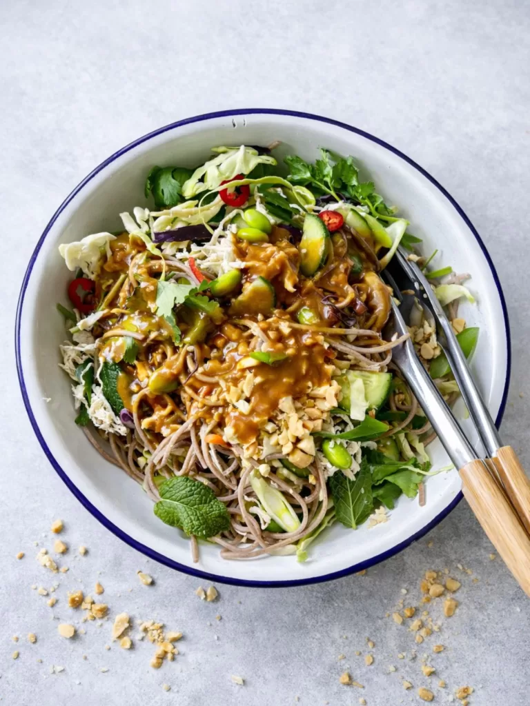Soba noodle salad with chicken and vegetables being tossed with creamy peanut dressing, topped with crushed peanuts and served in a bowl with salad tongs on a light grey background.