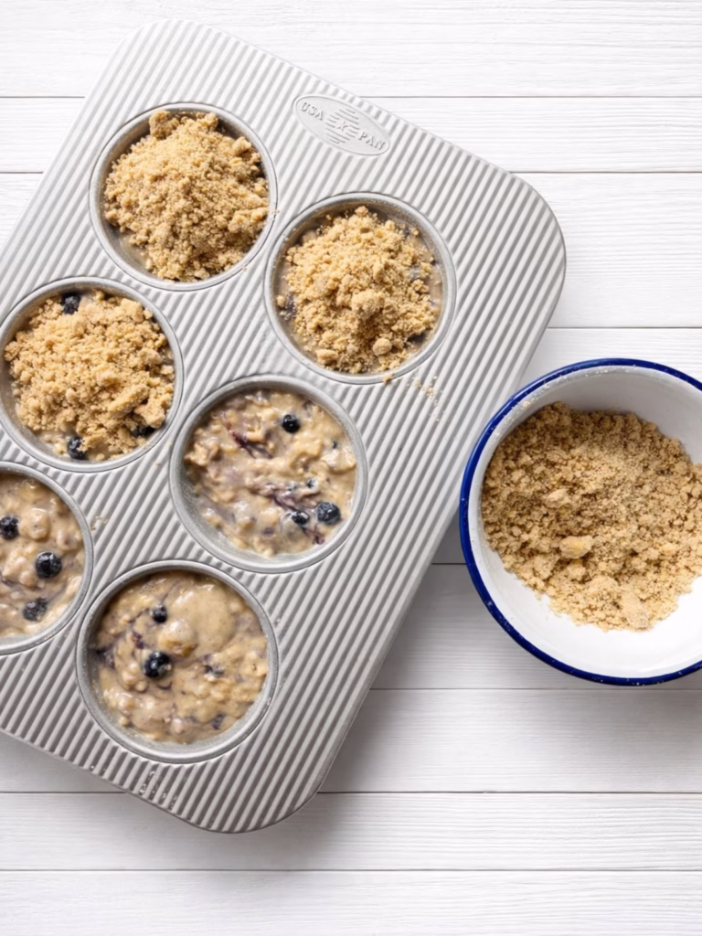 Blueberry muffin batter in a six hole muffin tin on a white timber surface, topped with crumb streusel, with a small bowl of extra crumble on the side