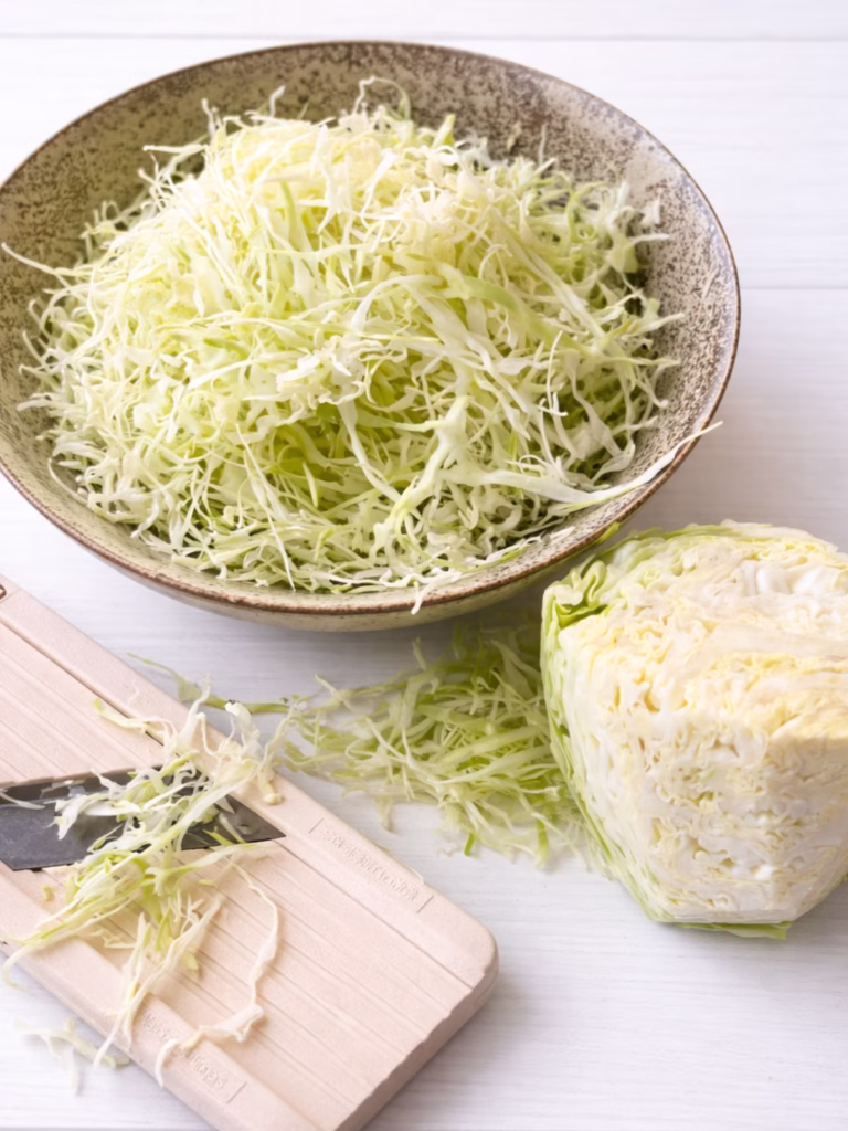 Finely shredded green cabbage in a bowl with mandolin slicer on a white timber background