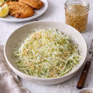 Sesame green cabbage slaw in a beige bowl topped with toasted sesame seeds, with crispy chicken schnitzel, lemon wedges, and parsley in the background on a white timber surface.