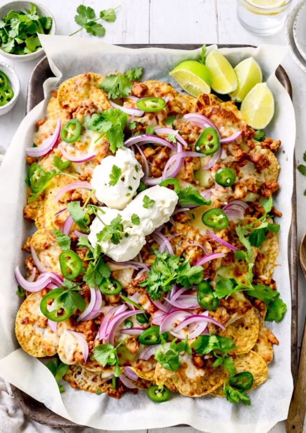Sticky chicken mince nachos on a tray with melted cheese, sour cream, jalapeños, red onion, coriander, and lime wedges on a white timber background