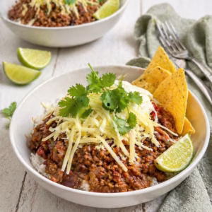 Bowl of chilli con carne served over rice with cheese, sour cream, coriander, lime wedges, and tortilla chips on a white timber background