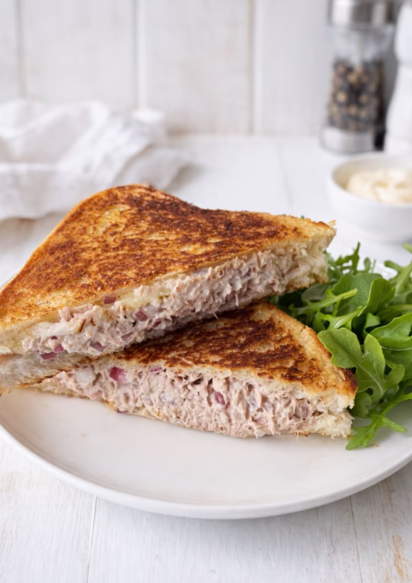 Tuna melt toastie cut in half on a white plate with rocket, showing a creamy tuna filling and golden toasted bread