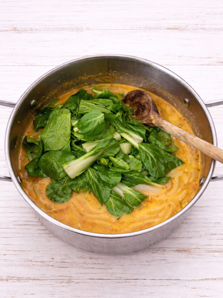 Fresh pak choy added to coconut curry udon in a pan, sitting on top of the sauce before wilting