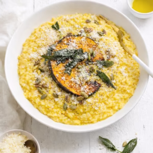 Overhead view of creamy pumpkin risotto in a white bowl topped with a roasted pumpkin wedge, crispy sage leaves, toasted pepitas, and grated parmesan on a white timber table with scattered sage and seeds.
