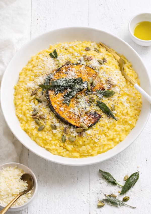 Overhead view of creamy pumpkin risotto in a white bowl topped with a roasted pumpkin wedge, crispy sage leaves, toasted pepitas, and grated parmesan on a white timber table with scattered sage and seeds.