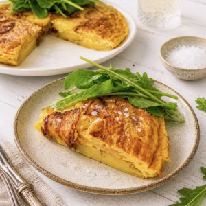 Slice of golden potato frittata on a ceramic plate with fresh rocket leaves, served on a rustic white timber table with the rest of the frittata in the background