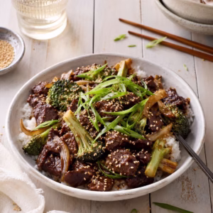 Chinese beef and broccoli stir fry served over fluffy white rice in a ceramic bowl, topped with sesame seeds and sliced spring onions. Styled on a white timber table with chopsticks