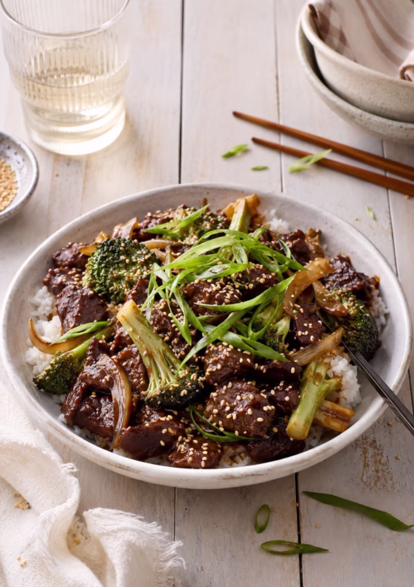 Chinese beef and broccoli stir fry served over fluffy white rice in a ceramic bowl, topped with sesame seeds and sliced spring onions. Styled on a white timber table with chopsticks