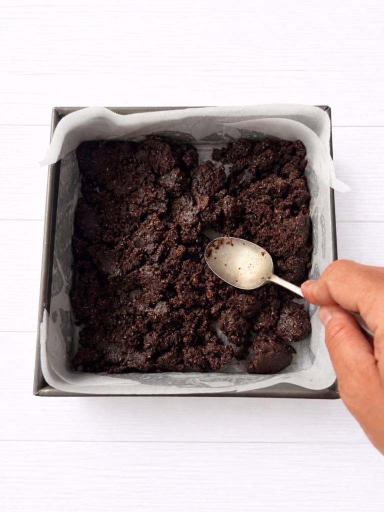 Chocolate base mixture being pressed into lined tin with a spoon ready for baking