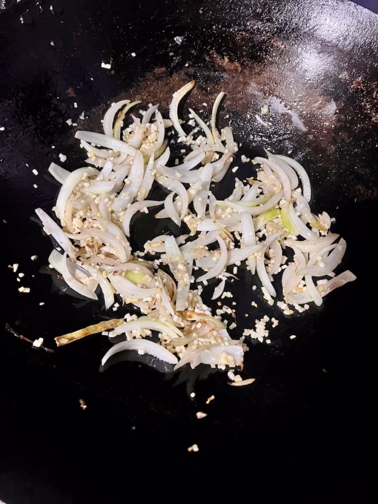 Close up image of sliced onion, garlic and ginger cooking in a black wok.