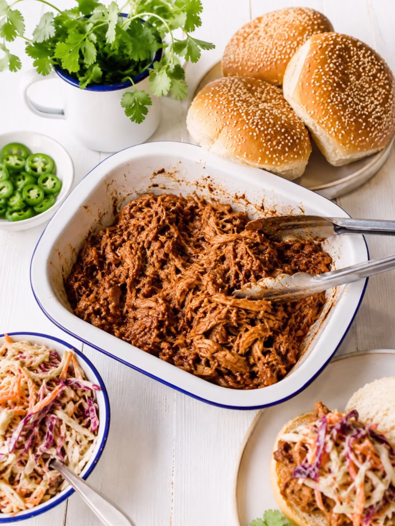 Pulled pork in a white enamel dish with sesame seed buns, coleslaw, jalapenos and coriander on a white timber background.