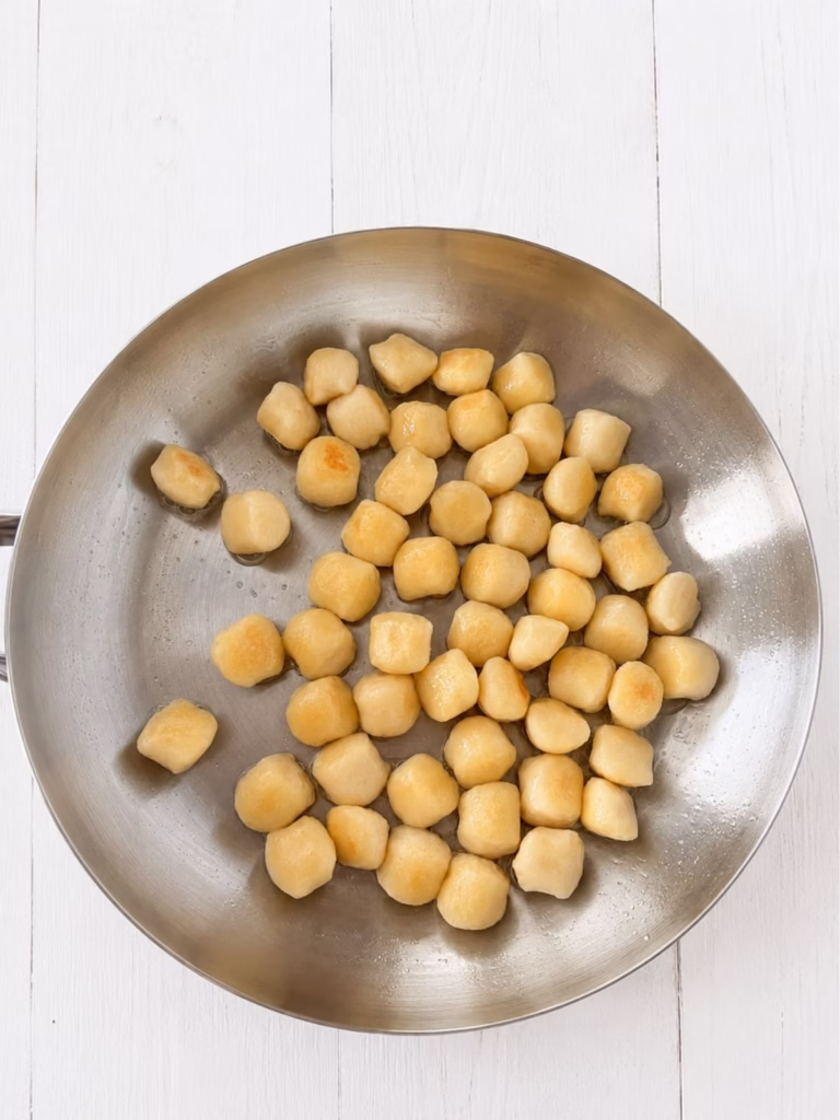 Raw gnocchi cooking in a stainless steel frying pan on a clean white timber background.