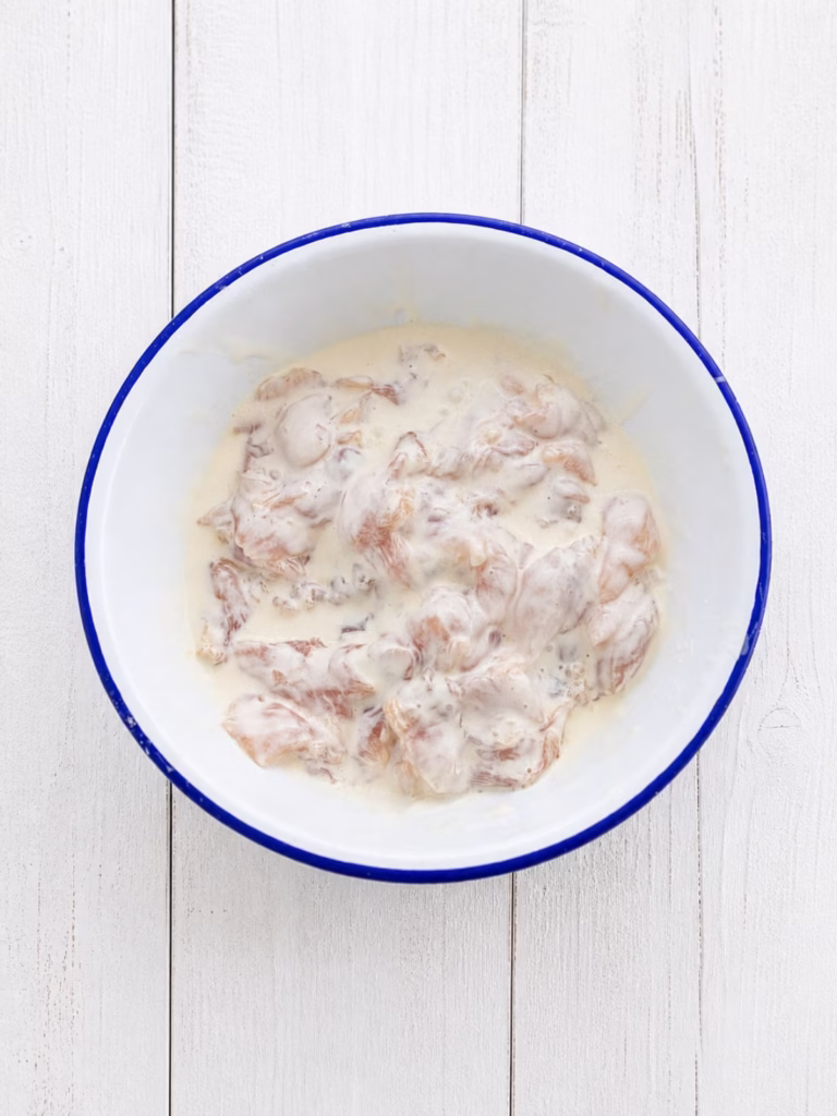 Thinly sliced chicken thigh fillets coated in batter in a bowl on a white timber background