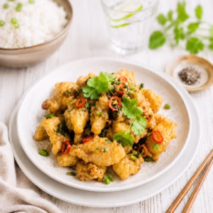 Crispy salt and pepper chicken served on a white plate with fresh chilli, coriander and spring onion, styled on a rustic white timber table with chopsticks, steamed rice and a lime water in soft natural light.