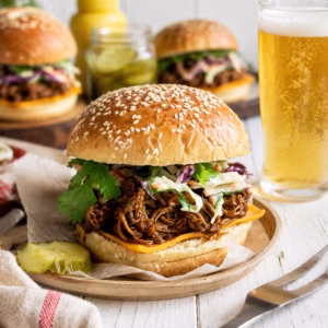 Slow cooker pulled pork burger with creamy coleslaw and melted cheese served with beer on a rustic white timber table.