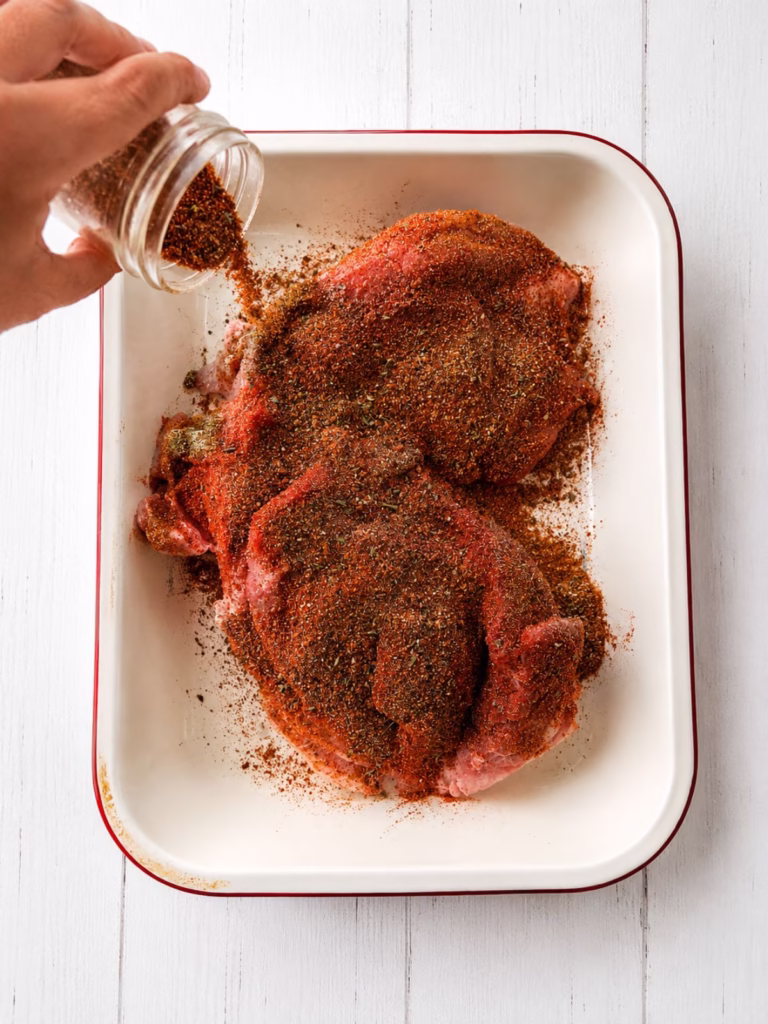 Spice rub being sprinkled over pork shoulder in a baking dish for slow cooker pulled pork.